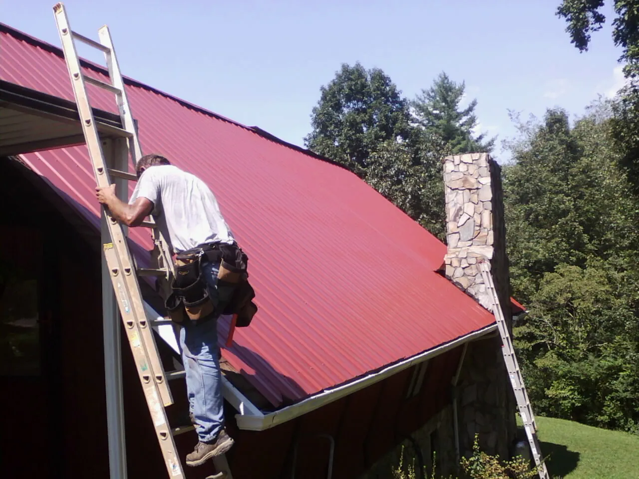 Roofer on a ladder beside a red metal roof and stone chimney — Soffit & Fascia Repair by Roofing Brothers in Christine
